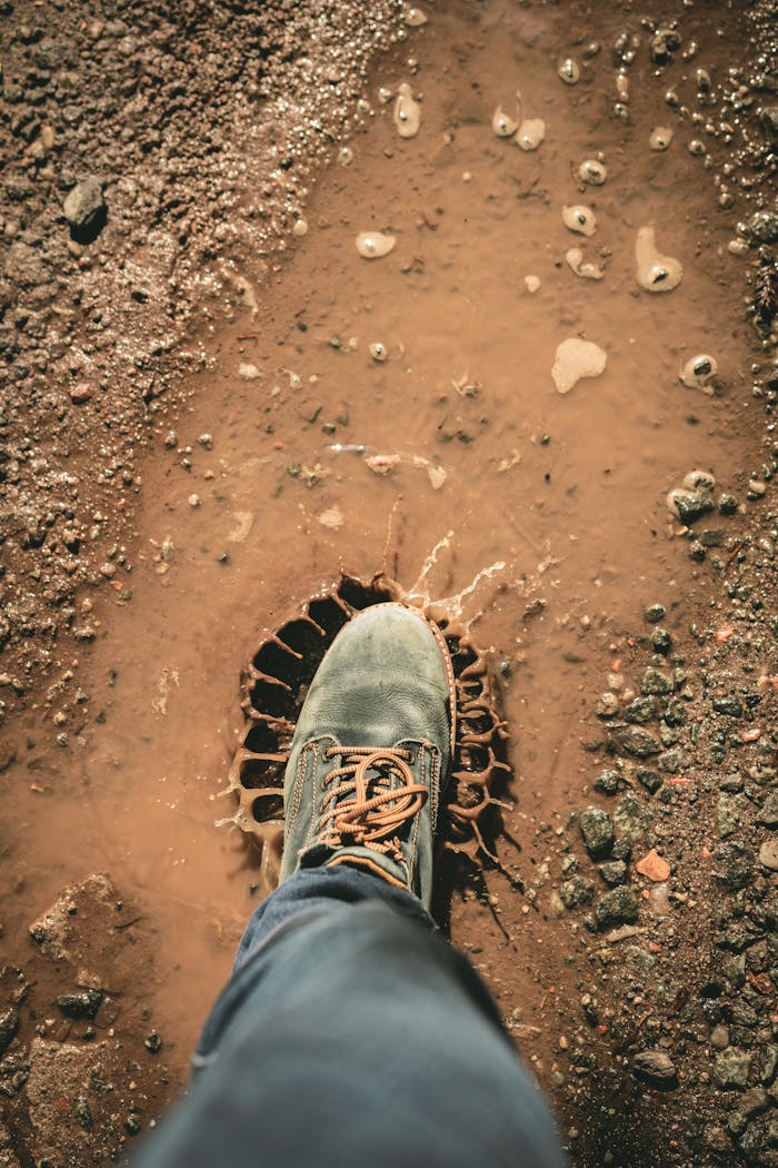A single boot stepping into a muddy puddle outdoors, creating a splash on a dirt road.