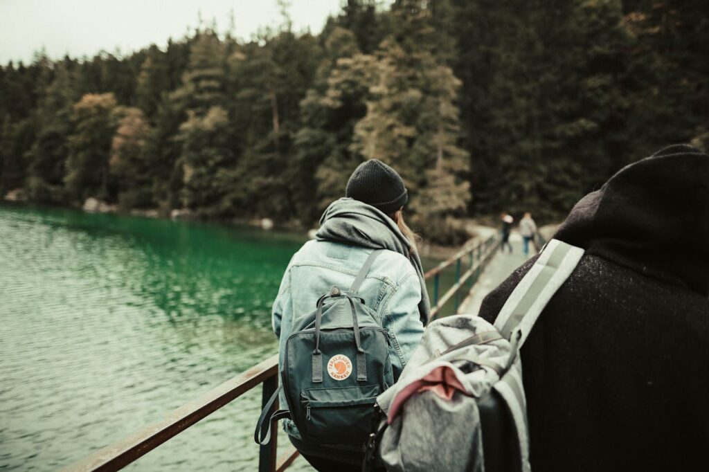 Two backpackers traverse a scenic wooden bridge amidst a lush forest and river backdrop in winter.